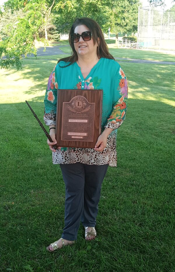 Standing on a lawn with trees in the background, a woman wearing a turquoise blouse, dark slacks, and sandals, holds a large rectangular wooden Lions Club plaque.
