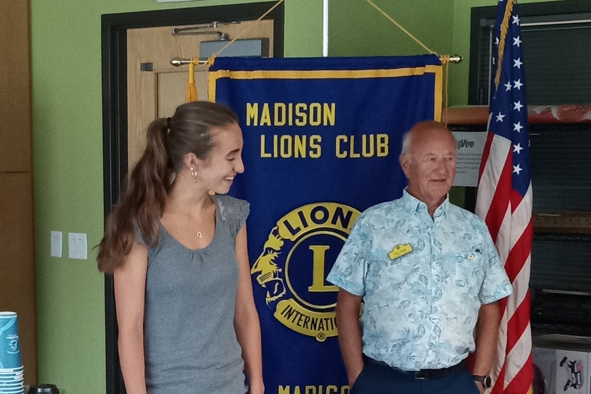 Standing in front of a banner reading Madison Lions Club, a man in a floral shirt presents a check to a young woman in a grey top and jeans